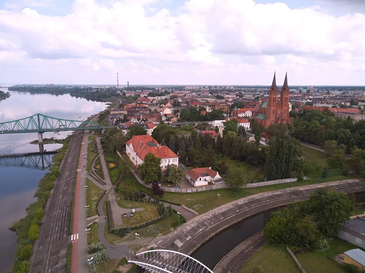 An aerial view of a city along a river, showing a green truss bridge, a large red-brick church with two spires, and numerous buildings with red roofs amidst green trees.