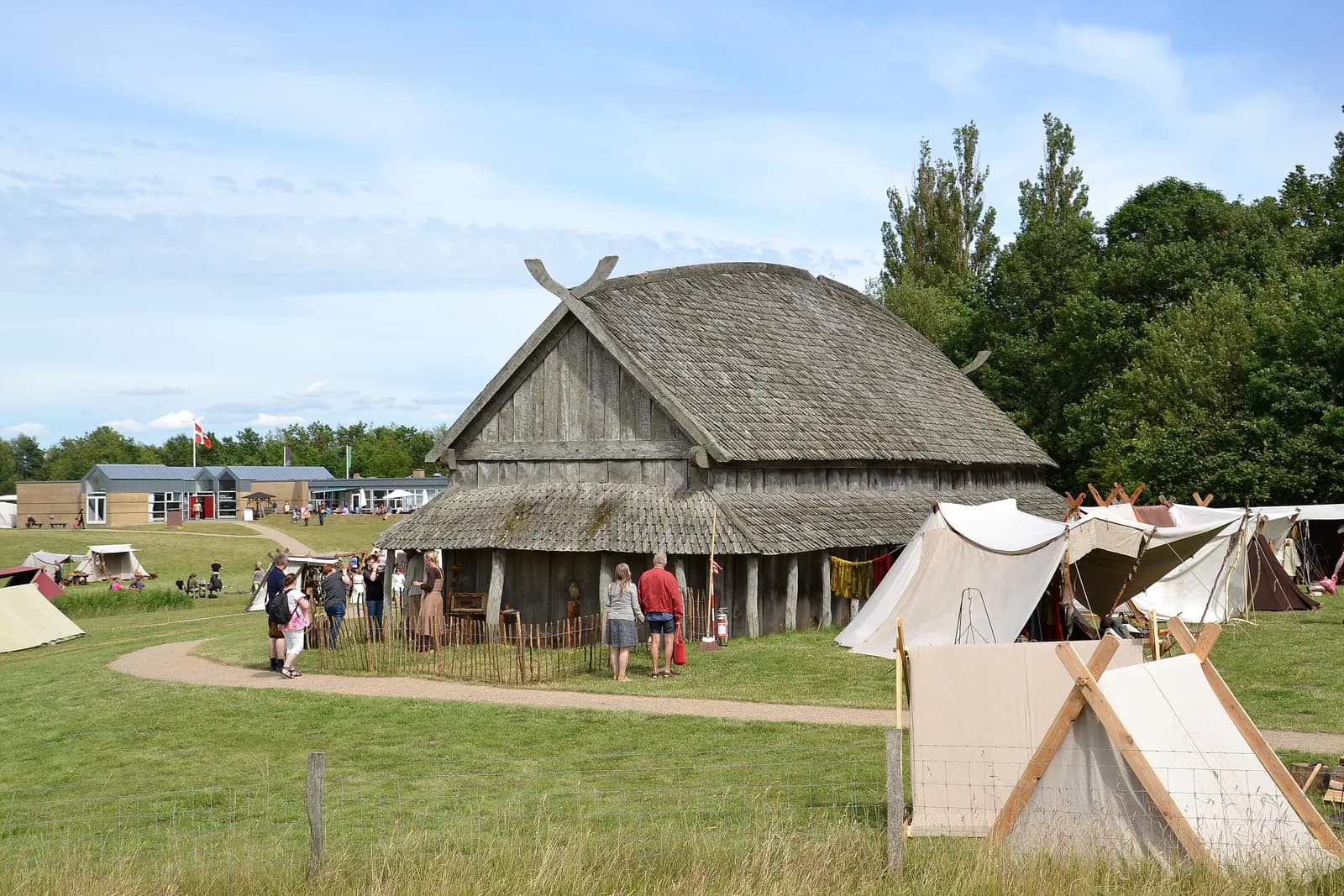 A large wooden building with a thatched roof, resembling an ancient longhouse, stands in a grassy field with several white tents and people walking around it under a blue sky.